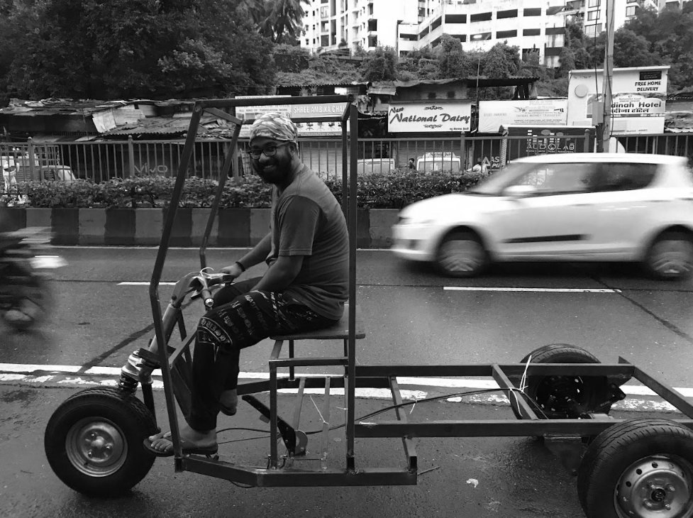 Black-and-white photo of the bare chassis being ridden on the road past traffic